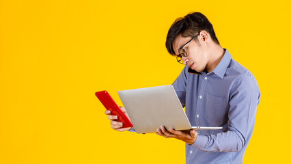 Studio shot of millennial busy stressed overload Asian professional male businessman employee holding laptop computer and tablet in hands on call with customer via smartphone on yellow background