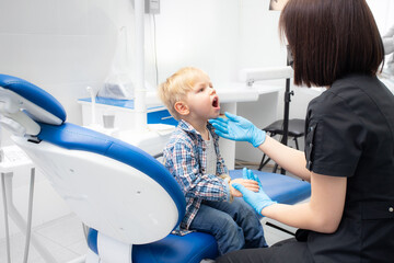 A cute little boy sits at the dentist, opened his mouth, showing his teeth to the dentist. Happy boy is sitting in the dentist's chair. Dentist - young brunette woman