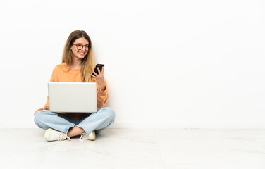 Young woman with a laptop sitting on the floor sending a message with the mobile