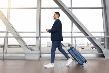 Young middle-eastern businessman with suitcase and smartphone in hands walking in airport