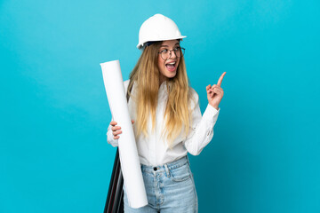 Young architect woman with helmet and holding blueprints isolated on blue background pointing up a...