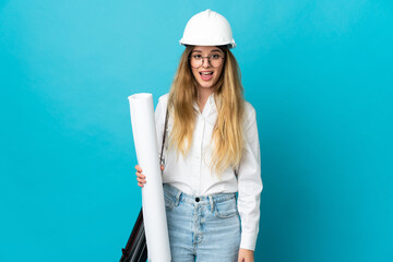 Young architect woman with helmet and holding blueprints isolated on blue background with surprise...