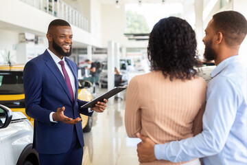 Smiling Black Salesman Consulting Family Couple In Car Dealership Center