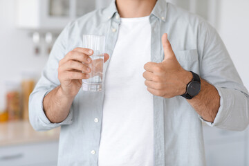 Unrecognizable adult caucasian man with glass of water showing thumb up sign on minimalist kitchen interior