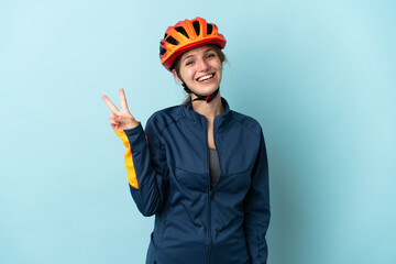Young cyclist woman isolated on blue background smiling and showing victory sign - obrazy, fototapety, plakaty