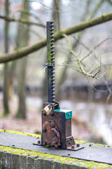 Old and rusty metal part with a cog gear in a floodgate on a bridge, fixed with a padlock so that it is not used, branches of trees and stream in a misty and blurred background in a nature reserve