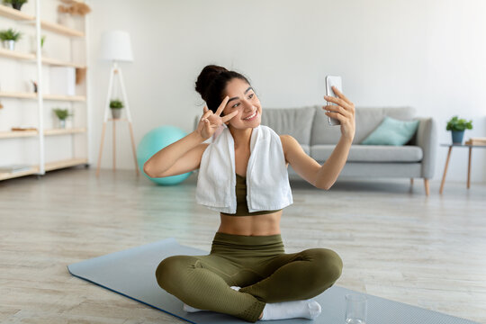 Happy Indian woman taking selfie during home workout, sitting on yoga mat indoors, full length