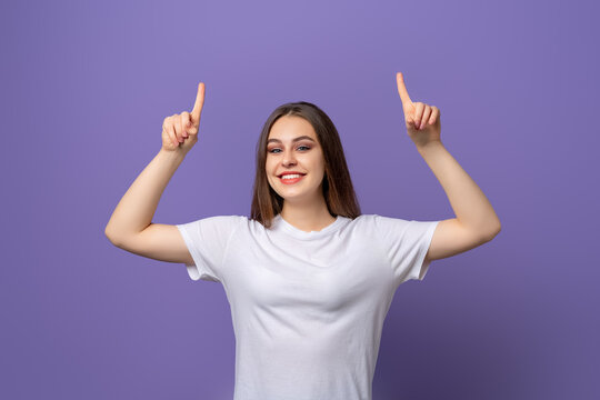 Smiling young woman with long hair pointing fingers up, raising eyebrows up surprised, showing advertisement, standing over purple background