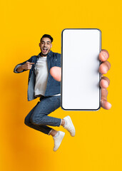 Young Arab guy jumping on air, pointing at cellphone with empty screen on orange studio background, mockup - obrazy, fototapety, plakaty