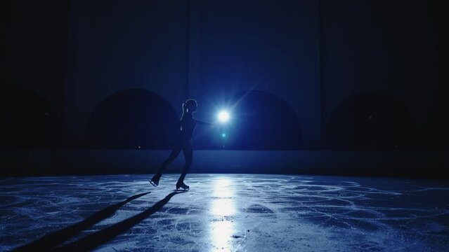 Blurred silhouette of figure skater girl is skating on ice arena in darkness, female figure is moving against blue soffit in slow motion. Full length shot, practicing sliding and spinning skills.