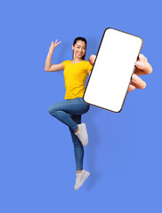 Full length of smiling young Asian woman jumping, gesturing peace and showing mobile phone with empty screen, mockup