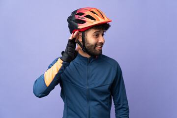 Young Moroccan cyclist man isolated on purple background listening to something by putting hand on the ear