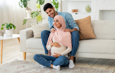 Pregnant muslim woman feeling relaxed, sitting on floor at living room and attentive husband massaging her shoulders