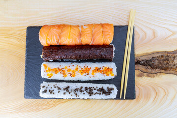 Sushi rolls of different kinds and chopsticks on a black plate on a wooden table.
