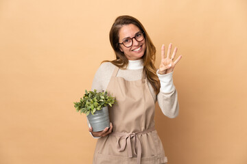 Middle age gardener woman holding a plant isolated on beige background happy and counting four with fingers