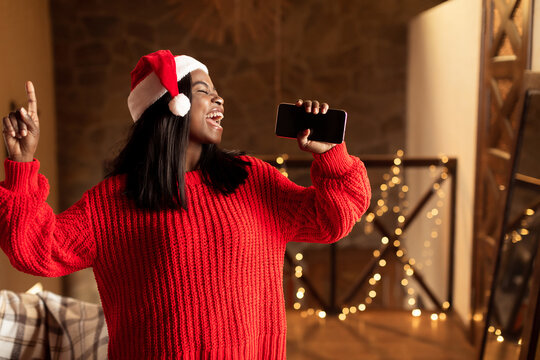 Christmas holiday music. Happy black lady in Santa hat singing, using smartphone as mic at home, mockup for mobile app