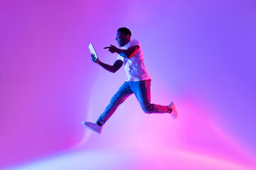 Fototapeta na wymiar Excited young black guy with tablet computer jumping in neon light, copy space. Cool app or advertisement concept