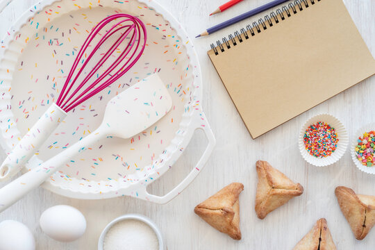 Baking background with silicone cake form, spatula and whisk, triangular cookies Hamantaschen, blank notebook paper on white wooden surface. Jewish holiday Purim baking background.