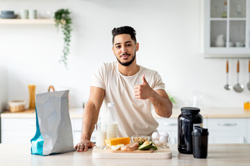Sporty young Arab guy with wholesome foods and protein shake gesturing thumb up at kitchen