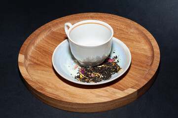 Dry tea leaves, scattered on a white saucer, a white ceramic cup on a wooden tray. black background