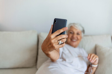 Portrait of happy cheerful beautiful elderly lady taking a selfie at home. Grandma taking selfies at home in the living room during the day. Senior woman sitting on sofa, taking selfie with smartphone