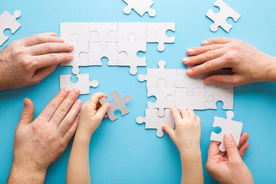 Mother, Father And Toddler Hands Playing And Assembling White Puzzle Pieces On Light Blue Table Background. Pastel Color. Family Activities. Spending Time Together. Closeup. Top Down View.