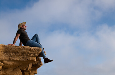 Fototapeta premium Adult man in cowboy hat sitting on cliff against sky. Almeria, Spain