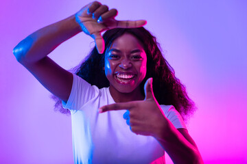 Happy black woman making picture frame with fingers, looking at camera and smiling for photo in neon light