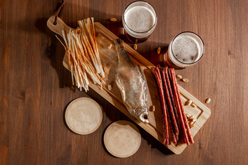 There are two full glasses of dark and light beer. On a wooden kitchen board there are cheese and meat sticks, and peanuts scattered around. 