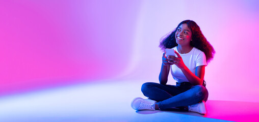 Amazing application. Young black lady using modern smartphone and looking at copy space in neon...