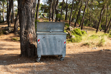 Fototapeta premium Old metal garbage container on the forest, trash dumpster. Dirty refuse bin. Garbage containers on the park. Recycling industry. Ecology, beautiful natural environment. Recycle bin front view.