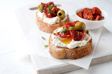 Two bruschettas with ricotta cheese, olive and dried tomatoes on ciabatta bread on white background. Olive oil and spices decorated. Copyspace.
