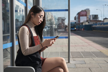 Young asian woman using her smartphone while waiting for the bus at bus stop.