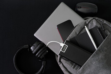 Silver laptop and modern gadgets lie in a gray backpack on a dark table. Travel, trip, study and...