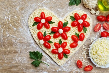 heart shaped pizza dough covered with mozzarella cheeses and flower shaped tomatoes,cauliflowers and parleys on wooden cutting board background.Love cooking concept for Valentine's day