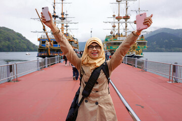 Portrait of smiling muslim woman on Togendai Station pier at Lake Ashi with Hakone sightseeing cruise pirate ship and mountain fog and cloudy sky background. Waving hands gesture and happy expression.