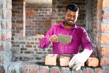 Happy smiling construction worker constructing or building wall by placing bricks and cement -...