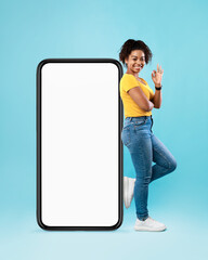 Cheerful black woman standing near big cellphone with empty white screen, gesturing okay on blue background, mockup