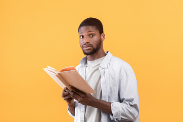 Obraz na płótnie Canvas Shocked black male student holding textbook, looking at camera with shock over yellow studio background
