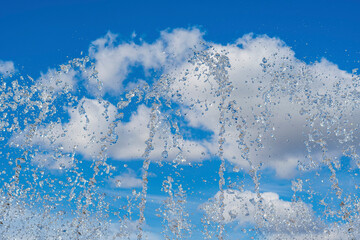A powerful jet of water rising up, drops and splashes against a blue cloudy summer sky. Refreshing fountain.