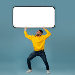 Arab guy holding huge heavy smartphone with empty white screen on blue studio background, mockup for ad