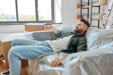 moving, people and real estate concept - tired man with boxes resting on sofa covered with plastic sheeting at new home