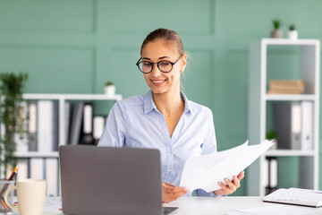 Busy female entrepreneur working on laptop and holding business reports, sitting at workplace in office interior