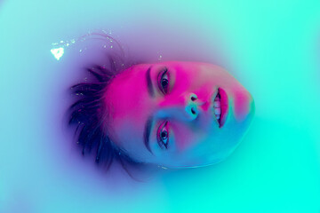 Top view of female face, young beautiful girl in milk bath with soft glowing in blue-green neon...