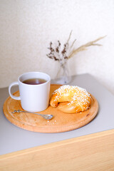 Bagel bun on a wooden round plate with a cup of tea and spoon. Cozy breakfast. Sweet organic plant-based traditional bakery. Modern beige neutral light food photo template.