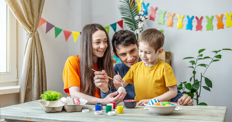 Fototapeta premium Mom, dad and little son in bright clothes are coloring eggs in a decorated room. Concept of family preparation for Easter, festive spring mood