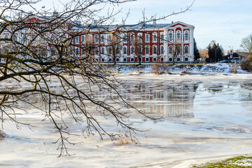 Fototapeta na wymiar Part of baroque Jelgava Palace is visible behind leafless winter branches and frozen icecovered river reflecting the building. Branches, winter, ice, river and baroque style university.