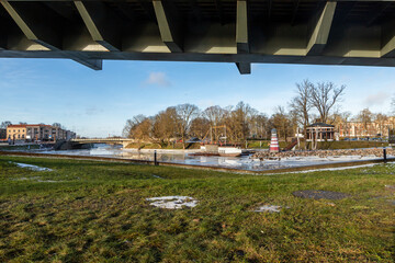 Obraz premium Low angle view under Mitava bridge to river Lielupe, Post Island, pavilion, palace, boat, lighthouse, riverbank, traffic bridge and hotel at winter. Sunny winter day under Mitava bridge in Jelgava.