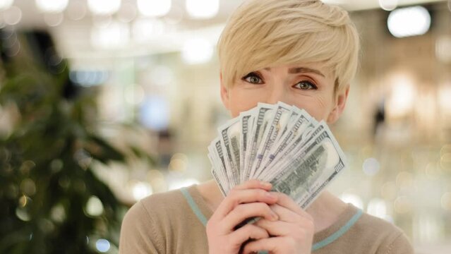Happy wealthy successful middle aged adult caucasian business woman looking at camera holding fan of money hiding behind dollar banknotes showing financial profit winning salary savings loan credit