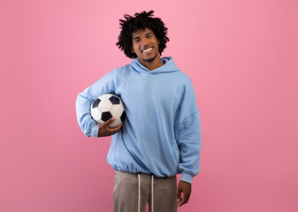 Portrait of positive black teenage guy holding soccer ball on pink studio background